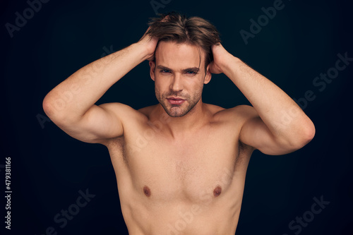 Handsome young bearded man isolated. Portrait of shirtless muscular man is standing on dark blue background and holding his hair