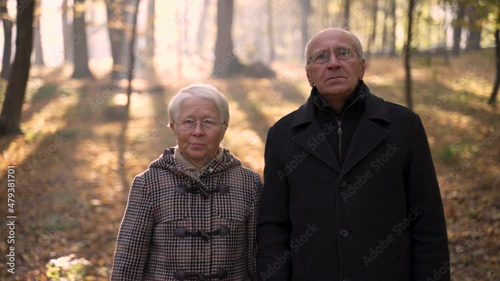 Elderly gray-haired woman and man posing for portrait with sad faces and then smiling while standing in the middle of forest park on sunny autumn day. Close-up of old pair spending time in nature Elderly gray-haired woman and man posing for portrait with sad faces and then smiling while standing in the middle of forest park on sunny autumn day. Close-up of old pair spending time in nature