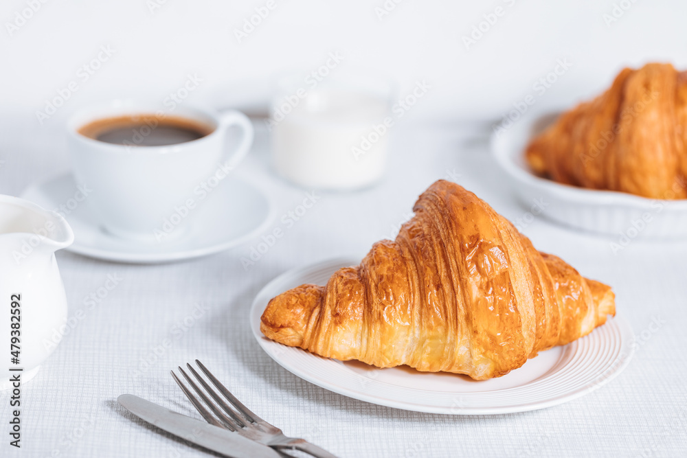 Croissant in bright white environment, with cup of coffee and milk and other croissant in blurry background. Simple and elegant breakfast setting and scene. Croissant in bright white environment, with cup of coffee and milk and other croissant in blurry background. Simple and elegant breakfast setting and scene.