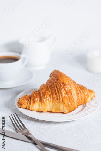 Croissant in bright white environment  with cup of coffee and milk and other croissant in blurry background. Simple and elegant breakfast setting and scene.