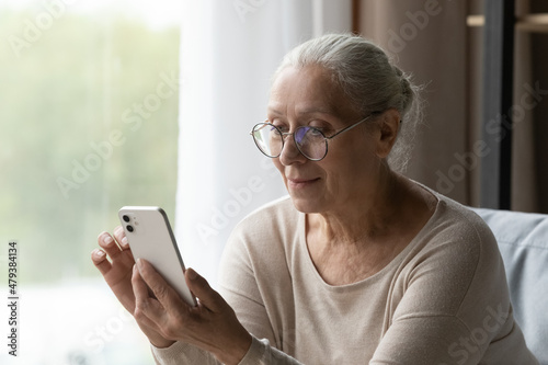 Happy middle aged old hoary retired woman in eyewear holding smartphone in hands, learning using software applications, typing message communicating with friends distantly or shopping online.