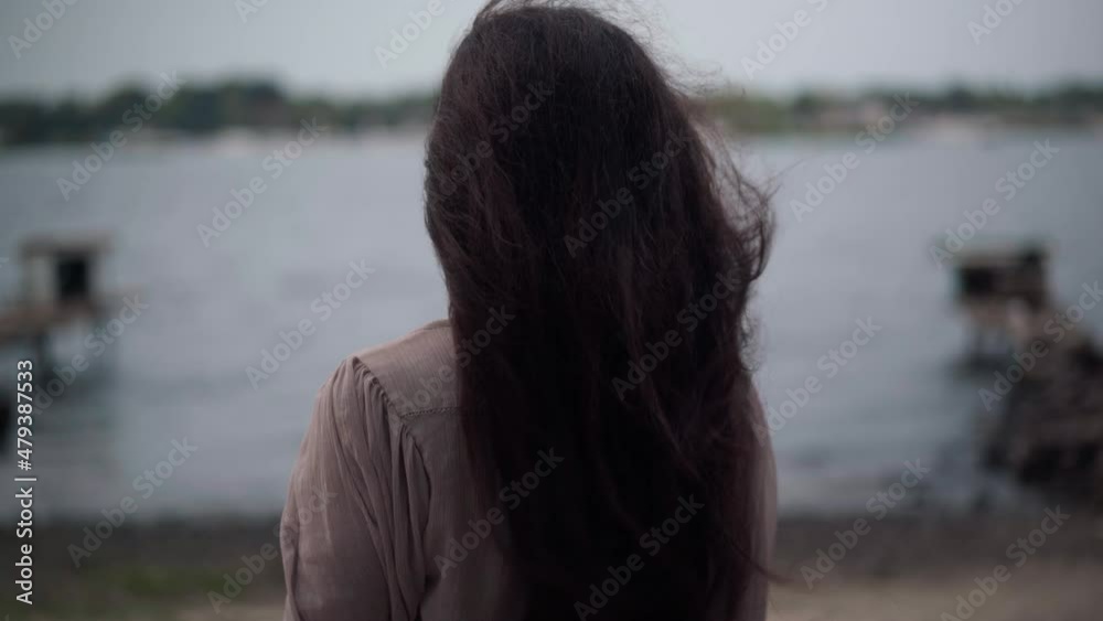 Back view senior woman with long hair blowing with wind standing at river bank looking away. Relaxed carefree happy Caucasian retiree enjoying cloudy day outdoors in parkland Back view senior woman with long hair blowing with wind standing at river bank looking away. Relaxed carefree happy Caucasian retiree enjoying cloudy day outdoors in parkland