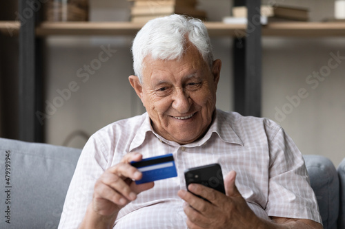Happy older senior hoary retired man holding plastic bank credit card and cellphone in hands, making payments in mobile shopping application, feeling satisfied with fast money transfer at home.