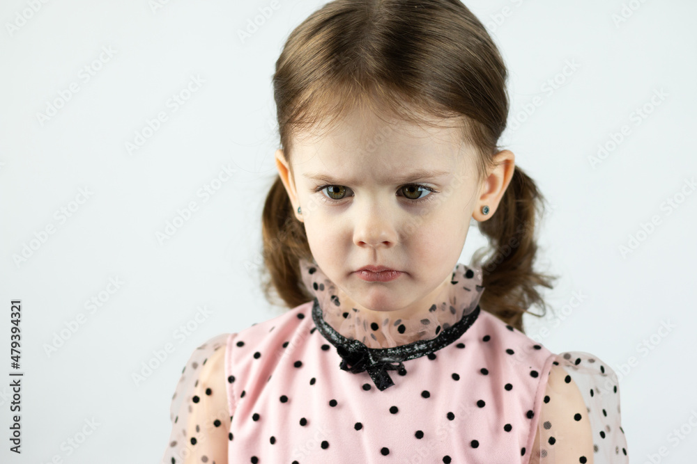 A little girl with two ponytails in a dress with polka dots on a white background looks down angrily. Studio photo A little girl with two ponytails in a dress with polka dots on a white background looks down angrily. Studio photo