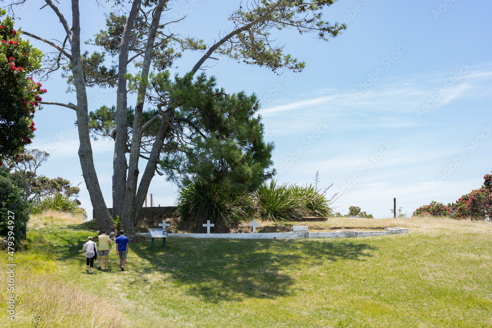 Auckland New Zealand -December 26 2021; Row of graves and crosses in white in shade of old pine tree and approaching tourists on Motuihe Island. Auckland New Zealand -December 26 2021; Row of graves and crosses in white in shade of old pine tree and approaching tourists on Motuihe Island.