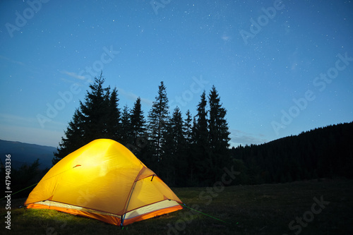 Brightly illuminated camping tent glowing on campsite in dark mountains under night stars covered sky. Active lifestyle and traveling concept