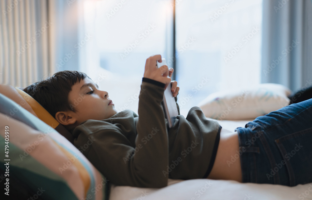 Happy Kid lying in bed holding tablet watching cartoon and chatting with friends on digital pad, Cute young boy playing games online on internet, Child relaxing at home with family Happy Kid lying in bed holding tablet watching cartoon and chatting with friends on digital pad, Cute young boy playing games online on internet, Child relaxing at home with family