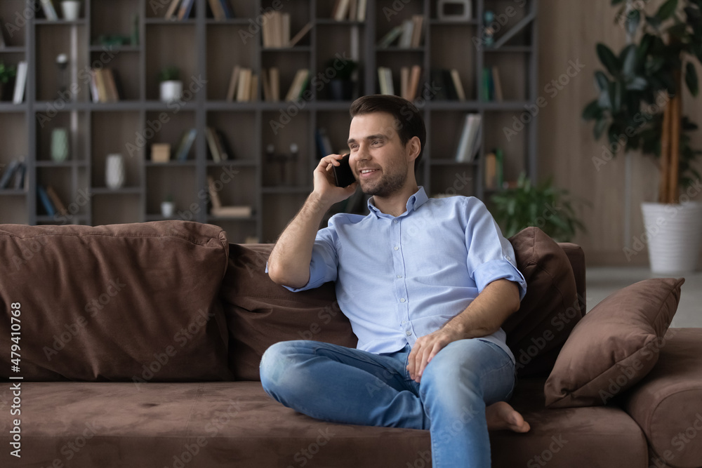 Smiling man chatting by phone call, relaxing on comfortable couch in modern living room, happy young male speaking, enjoying pleasant conversation with friend or girlfriend by smartphone at home