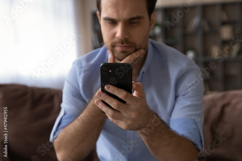 Close up thoughtful man looking at smartphone screen, touching chin, sitting on couch, pensive businessman thinking, reading news in email or message, hesitating and doubting, pondering offer