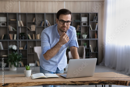 Portrait of thoughtful businessman in glasses standing near work desk with laptop in modern cabinet, home office, touching chin, looking at camera, confident entrepreneur freelancer looking at camera