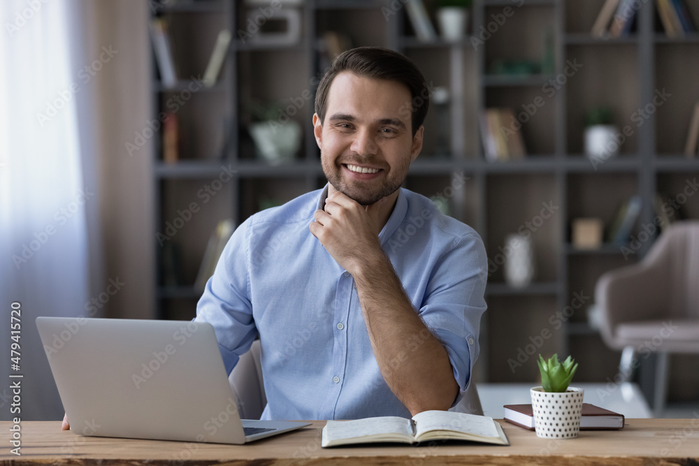 Head shot portrait of smiling successful businessman sitting at workplace desk with laptop, happy young man freelancer or student looking at camera, working or studying online, home office