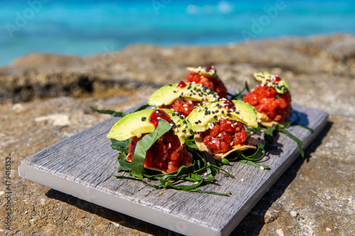 Fresh avocado and tuna fish on Mexican tostada for lunch. Food is on a wooden board on a beach with tropical turquoise ocean water in the background. photo