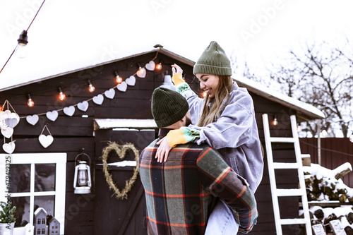 couple of young woman and man having fun in courtyard of decorated suburban house in winter, concept of Christmas and New Year vacation on farm, family love and support, Valentines Day celebration