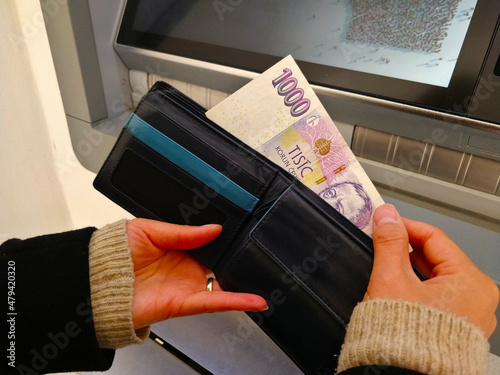 Someone puts paper money out of the wallet in front of the ATM. The wallet is black. The banknote is one thousand Czech crown.
