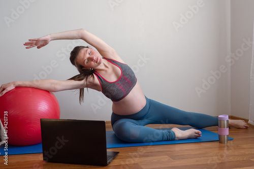 Fitness pregnant woman doing yoga workout on yoga mat opposite laptop with online master class. Healthy sports pregnancy concept.  photo