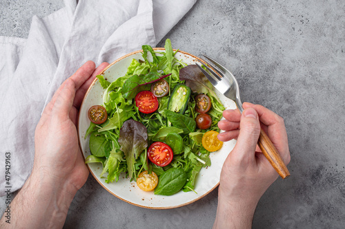 Man eating vegetarian vegetables healthy salad with red and yellow cherry tomatoes and green salad leafs on white plate on gray concrete background top view, healthy food and diet concept