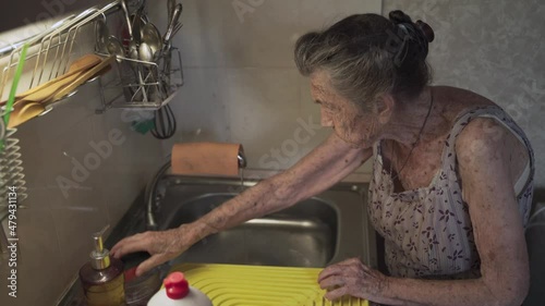 Senior elderly woman washing dishes in kitchen. Sad mature housewife cleans up the old dishes in the kitchen. Upset woman with gray hair 90 years old washing kitchen utensils in a hand wash at home.