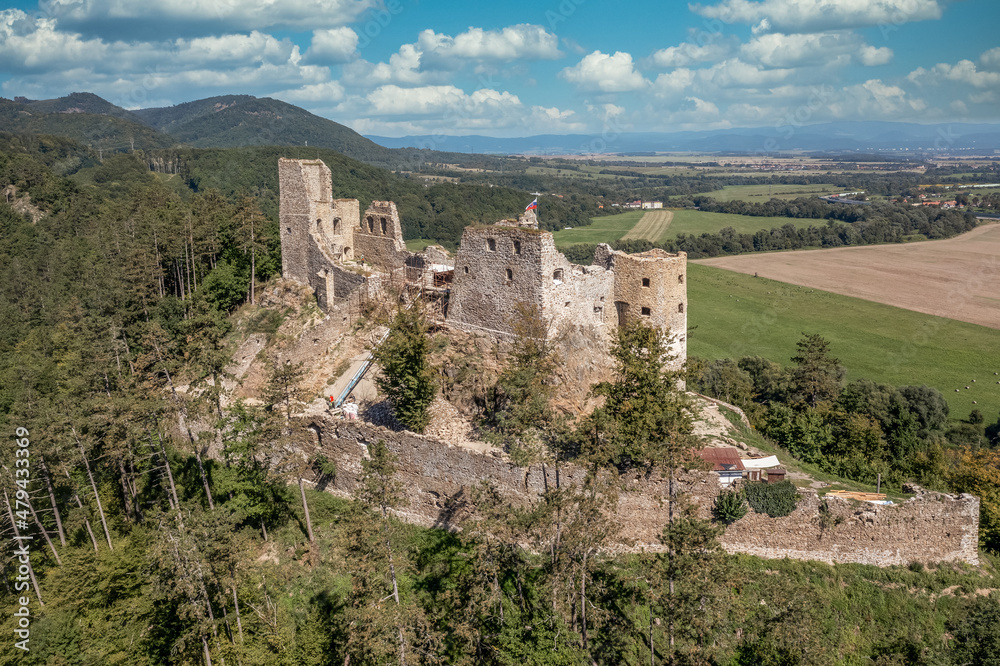 Aerial view of under restoration medieval Reviste castle above the Hron (Garam) river in Slovakia with donjon, circular gate tower, ruined gothic palace blue cloudy sky 