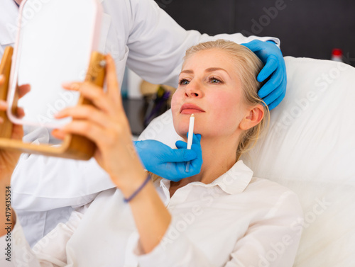 Young woman sitting in armchair and holding mirror while cosmetologist using special dermatograph pencil for marking skin.