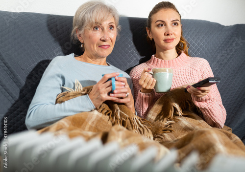 Senior woman and her adult daughter sitting on sofa covered with plaid, drinking hot tea and trying to get warm by using radiator in living room.
