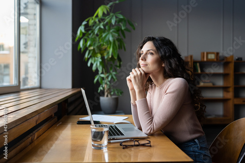 Young dreamy smiling female entrepreneur thinking about business goals and objectives, enjoying good window view while working in office, pleased businesswoman resting from computer work photo