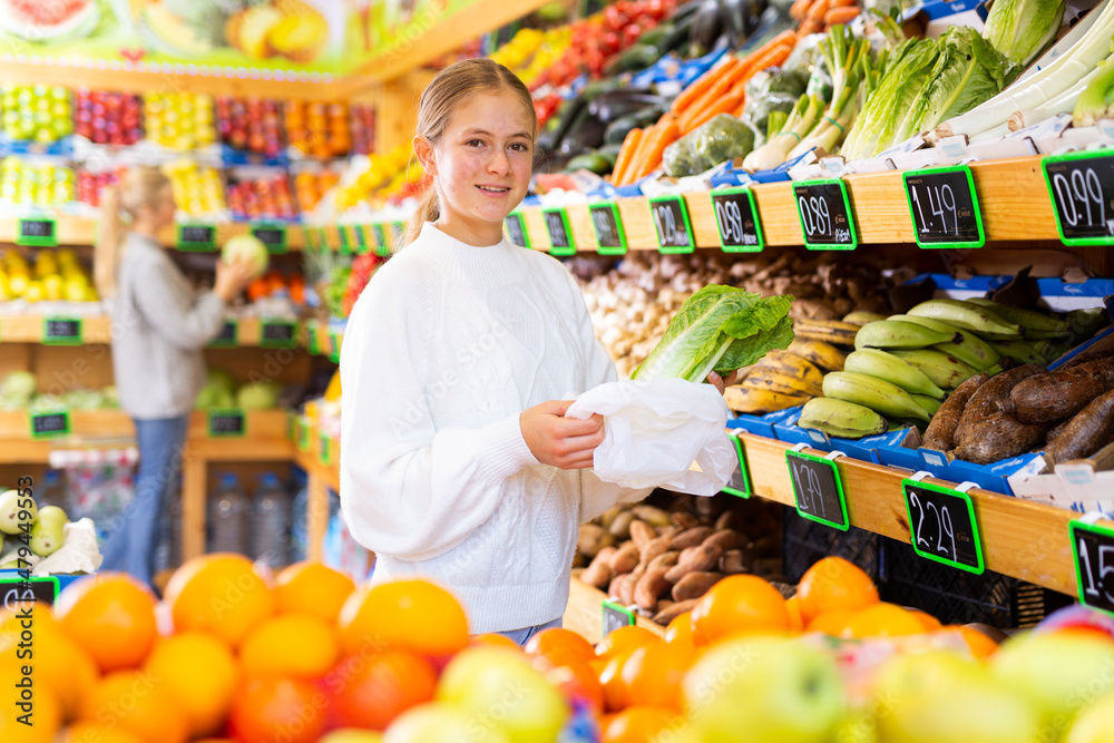 Portrait of smiling cute teen girl choosing fresh organic romaine lettuce in fruit and vegetable department of supermarket