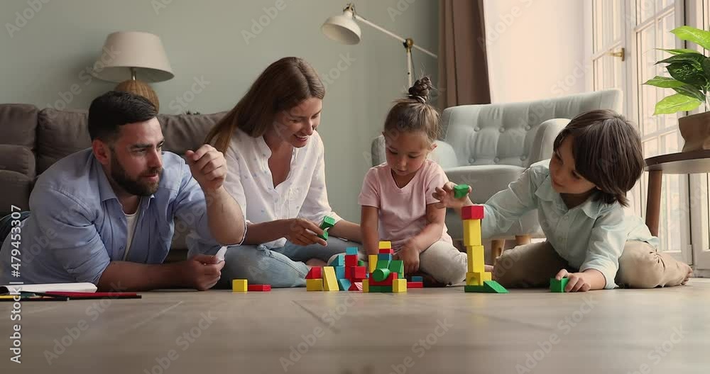 Smiling happy parents with small daughter son gather together on warm heated floor play creative game. Foster mom dad two adopted kids construct fantasy buildings from wood blocks at cozy living room