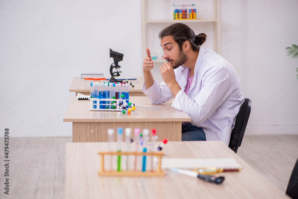 Young male chemist sitting at the desk in the classroom