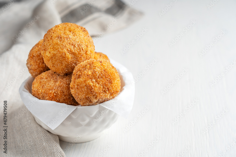 Italian homemade Arancini rice balls or croquettes stuffed, coated with bread crumbs and deep fried served in bowl as appetizer on white wooden background with textile towel. Image with copy space