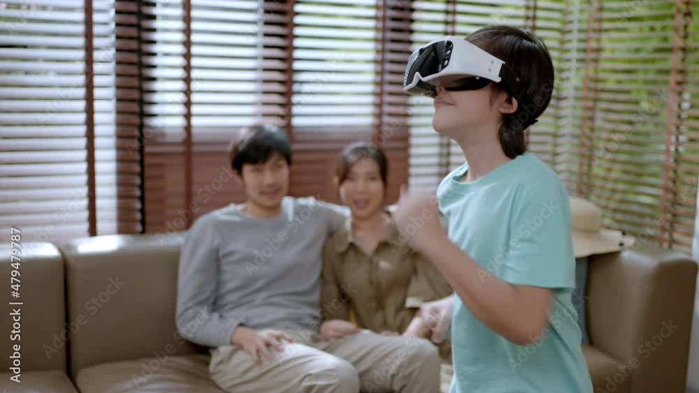 Technology and Family concept.Happy asian family at home on living room sofa having fun.They are looking boy playing games using virtual reality headset(VR) at home. Technology and Family concept.Happy asian family at home on living room sofa having fun.They are looking boy playing games using virtual reality headset(VR) at home.