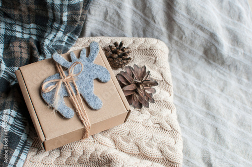 A gift cardboard box tied with jute and decorated with a gray felt figurine of an elk or deer lies on a light sweater with braids, fir cones, checkered fabric. Cozy zero waste holiday