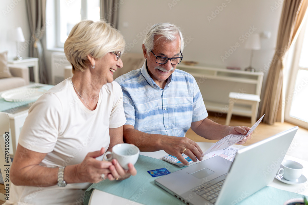Obraz premium Happy middle aged husband and wife sitting at table with laptop and paper bills, calculating domestic incomes together at home.