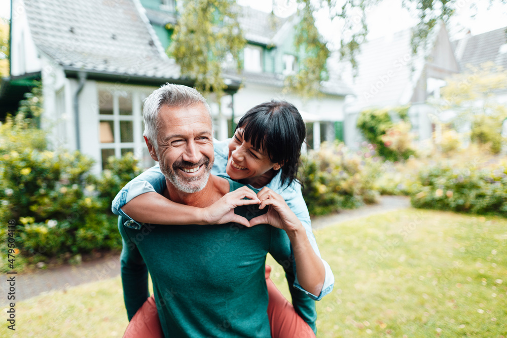 Cheerful man giving piggyback ride to woman gesturing heart shape in backyard Cheerful man giving piggyback ride to woman gesturing heart shape in backyard