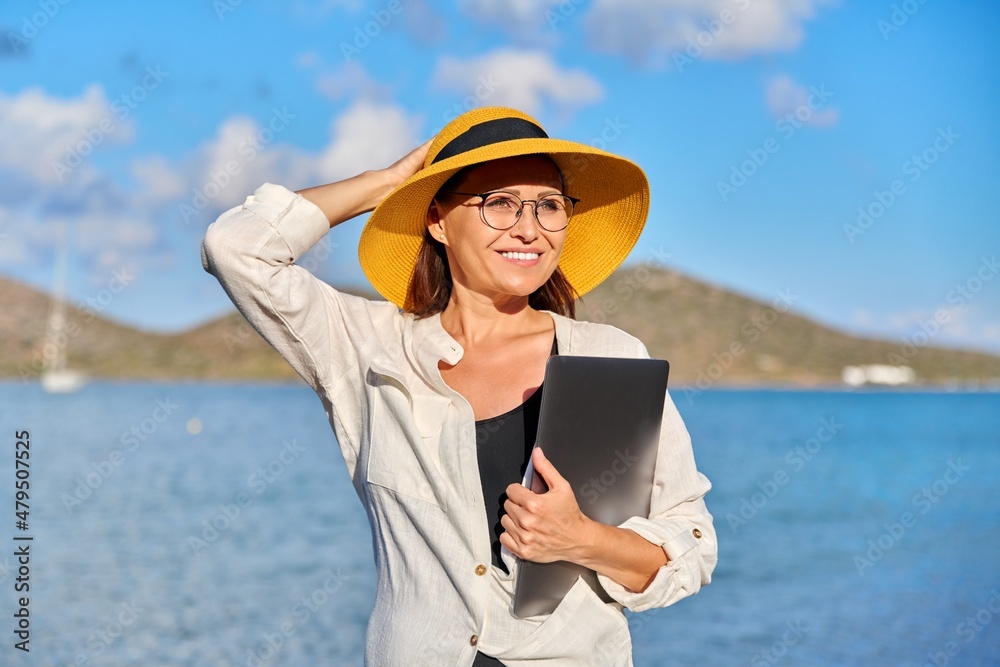 Outdoor portrait of mature woman in hat on beach with a laptop in hands