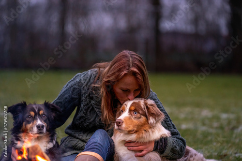 Young woman is sitting outside in the woods with her two Australian Shepherd dogs. Snow on the grass, twilight by the campfire. Cuddles and companion animals
