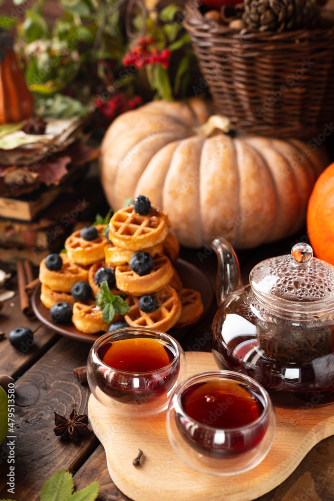 many waffles are stacked on a plate, decorated with mint leaves and blueberries. Tea in a glass teapot and two small bowls