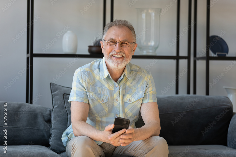 Portrait of smiling positive old senior man holding cellphone in hands, shopping online, communicating in social network, using software applications, relaxing on comfortable sofa alone at home.