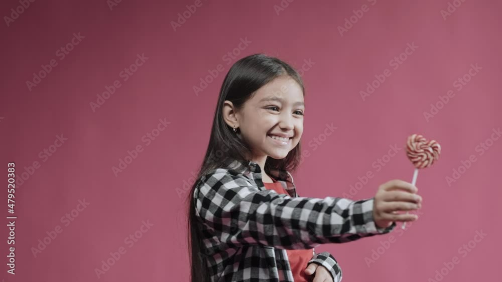 Little girl is happy with heart caramel on a stick, surprised emotion. Cute kid smiling holding candy in the studio with pink background. High quality 4k footage Little girl is happy with heart caramel on a stick, surprised emotion. Cute kid smiling holding candy in the studio with pink background. High quality 4k footage