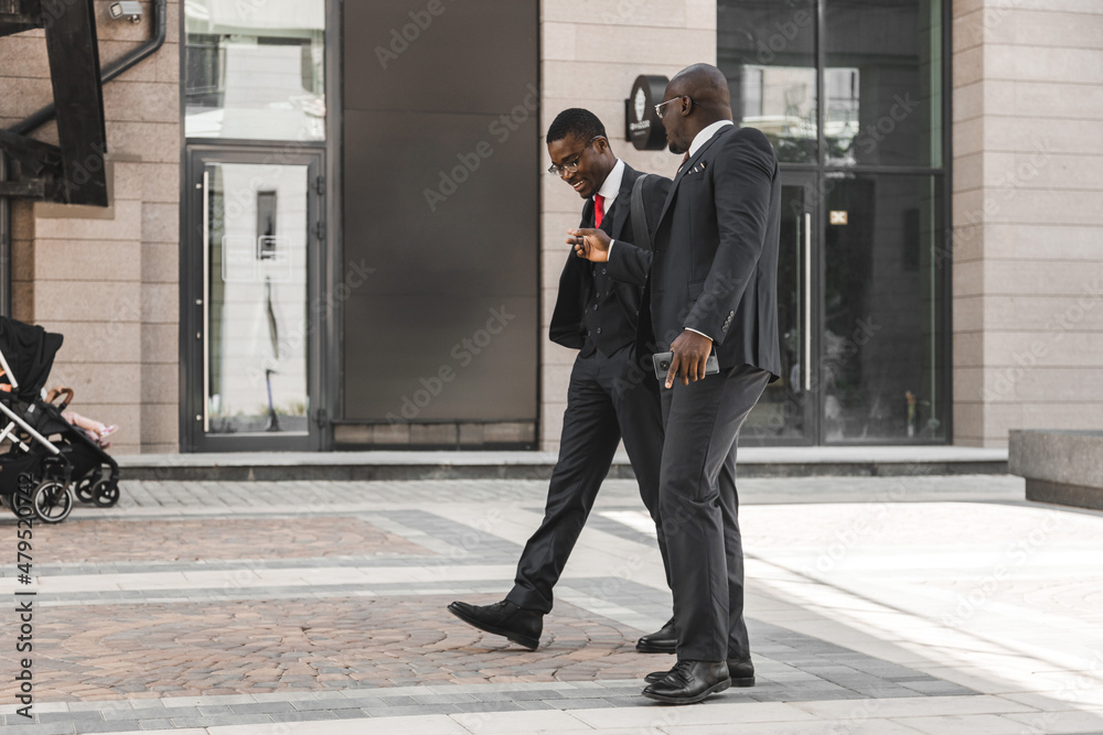Two dark-skinned african american businessmen in suits and glasses walk outdoors in the city and discuss business