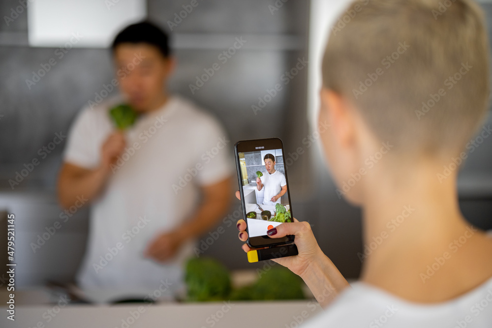 Fototapeta premium Woman taking video on smartphone of asian man cooking vegetarian salad on kitchen. Concept of video blogging. Idea of healthy and diet eating. Adult male blogger with grapes looking at mobile phone
