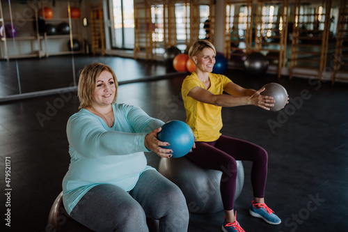 Happy overweight woman sitting on fintess ball and exercising with personal trainer in gym photo