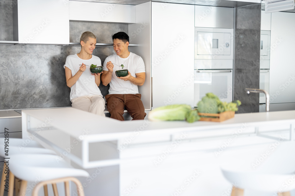 Laughing multiracial couple eating salad on kitchen at home. Concept of healthy and diet eating. Idea of domestic lifestyle. Wide interior view with copy space Laughing multiracial couple eating salad on kitchen at home. Concept of healthy and diet eating. Idea of domestic lifestyle. Wide interior view with copy space