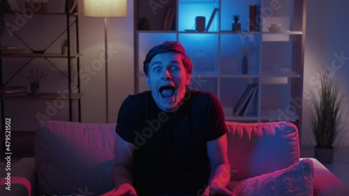 Grimacing man. Meme expression. Silly behavior. Foolish guy making nuts scare face sitting sofa in dark neon light home interior looped. photo