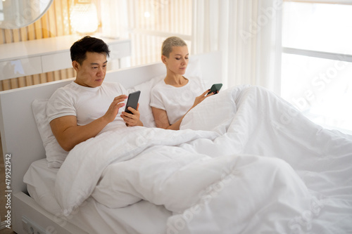 Asian man showing something on smartphone to his caucasian girlfriend. Concept of relationship and spending time together. Smiling multiracial couple lying under blanket in bed. Bedroom interior