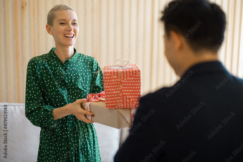 Smiling asian man giving gift present to his girlfriend. Concept of holiday, event and birthday. Idea of family relationship. Back view of woman wearing green dress. Interior of modern apartment Smiling asian man giving gift present to his girlfriend. Concept of holiday, event and birthday. Idea of family relationship. Back view of woman wearing green dress. Interior of modern apartment