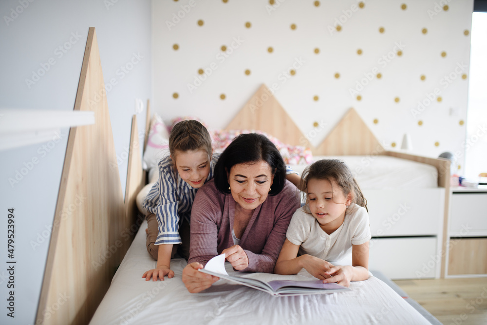 Happy small girls with senior grandmother indoors in bedroom at home, reading on bed.
