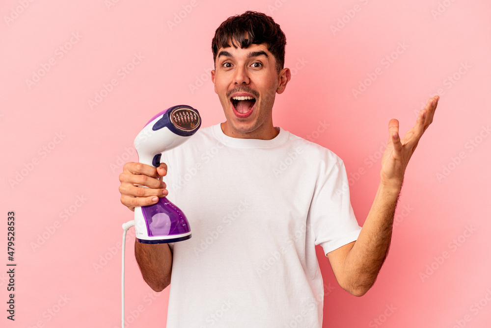 Young mixed race man holding an iron isolated on pink background receiving a pleasant surprise, excited and raising hands. Young mixed race man holding an iron isolated on pink background receiving a pleasant surprise, excited and raising hands.