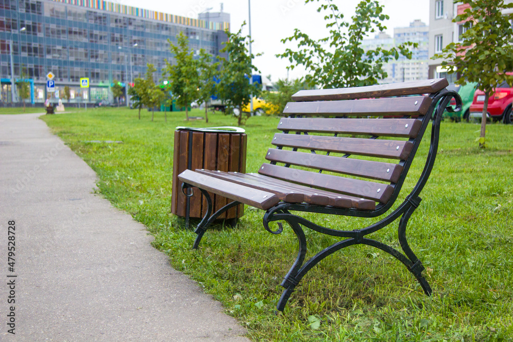 A wrought iron bench and an urn of wooden planks on an alley, avenue, a walking street against the backdrop of a modern city, a shopping center and cars. A wrought iron bench and an urn of wooden planks on an alley, avenue, a walking street against the backdrop of a modern city, a shopping center and cars.