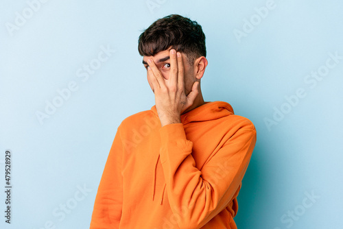 Young mixed race man isolated on blue background blink at the camera through fingers, embarrassed covering face. © Asier