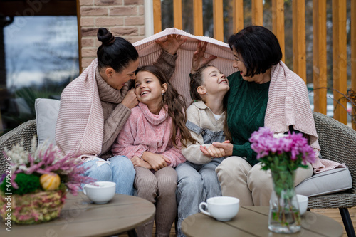 Two happy sisters with mother and grandmother sitting wrapped in blanket outdoors in patio in autumn